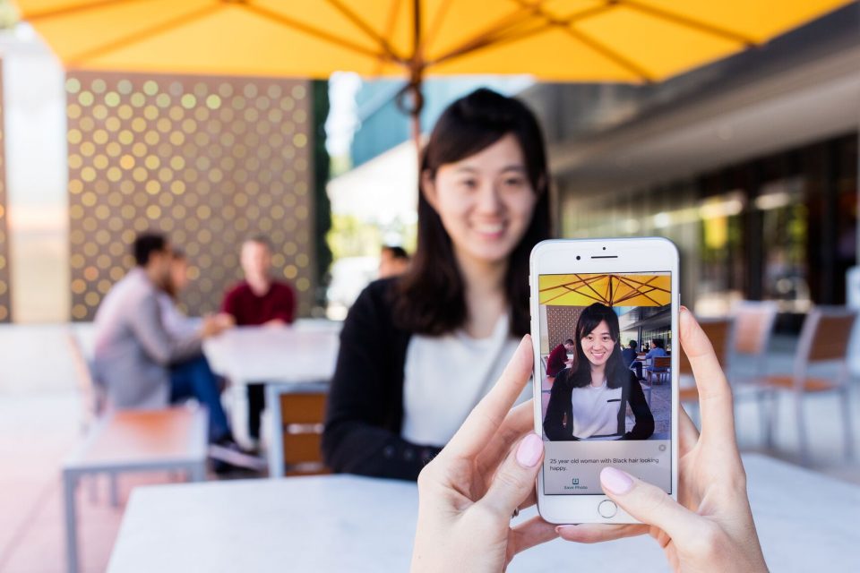 A photo of Seeing AI on an iPhone pointing at a woman with black hair. Seeing AI describes the woann as a "25 year old woman with black hair looking happy."