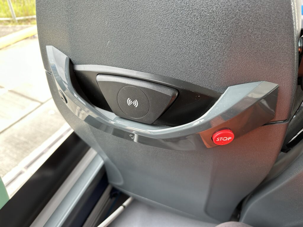 Back of a Bus seat showing a wireless chargine slot, USB A port for chargine and a stop button with a braille S. Slightly out of view is a downward facing lamp and two bag hooks.