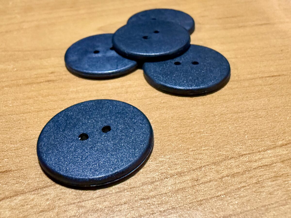 An image of five NFC Laundry Buttons. Four are in a pile with one by itself. All are black and, on a desk, wooden desk.